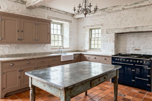 A light-filled French Country kitchen features whitewashed stone walls, distressed ceiling beams, and two sash windows painted sage green. Warm taupe Shaker cabinets with polished nickel latches support pale marble counters and a porcelain farmhouse sink; a weathered zinc-topped worktable stands on terracotta tiles, and a glossy navy, vintage-style range sits within a stone alcove beneath a rustic mantel and metal candelabra chandelier.