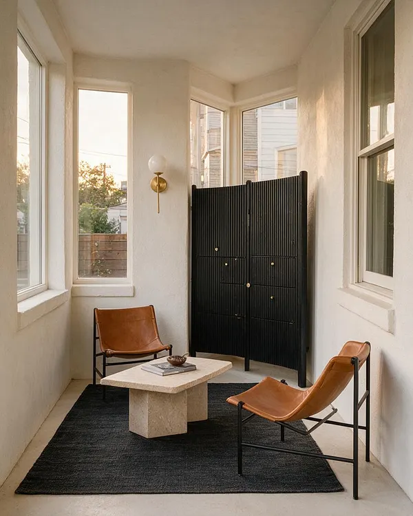 A narrow modern front porch with white stucco walls, tall windows, and a black ribbed folding screen at the far end. Two tan leather sling chairs sit on a dark rug around a light stone coffee table, with a brass-and-glass wall sconce adding warmth.