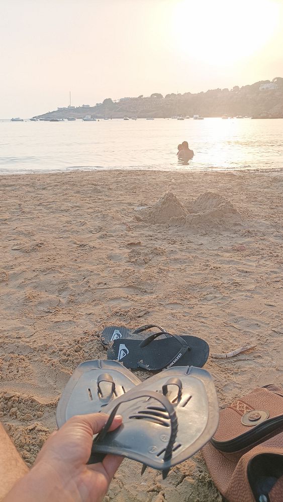 A person is preparing to go swimming at the beach, holding flip-flops in hand.