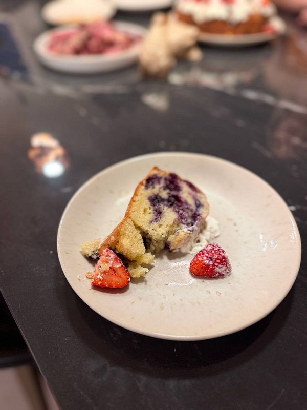 A partially eaten slice of blueberry cake with powdered strawberries on a white plate on a dark countertop.