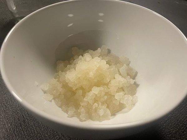 Close-up image of a kefir SCOBY floating in a cloudy liquid inside a round glass container on a wooden surface.