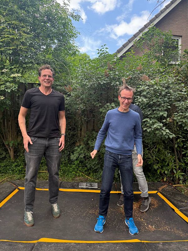 Two men are jumping on a trampoline in a backyard setting.