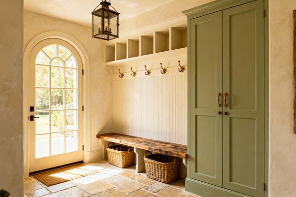 A sunlit mudroom features an arched, multi-pane French door beside a built-in bench with a rustic wood seat, white beadboard back, and brass double hooks beneath open cubbies. A tall sage-green cabinet with antique brass pulls stands to the right, woven baskets tuck under the bench, and tumbled limestone tiles, creamy plaster walls, a coir mat, and a glass-and-iron lantern pendant complete the relaxed French Country look.