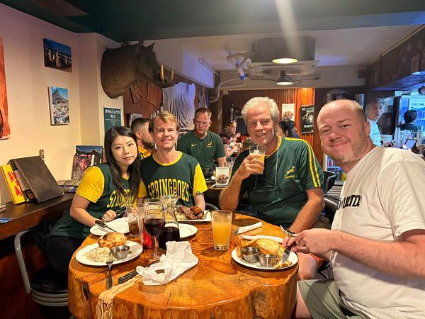 A group of four people sit at a round wooden table in a South African-themed pub, enjoying food and drinks while watching a sports match.