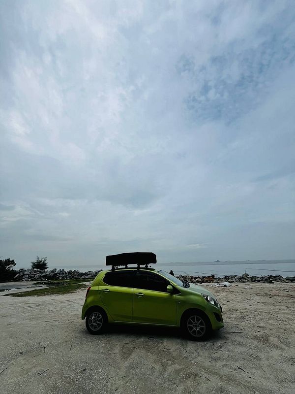 A woman in a black swimsuit floats on her back in crystal clear blue ocean water near a rocky shore with a cliff and cloudy sky in the background.