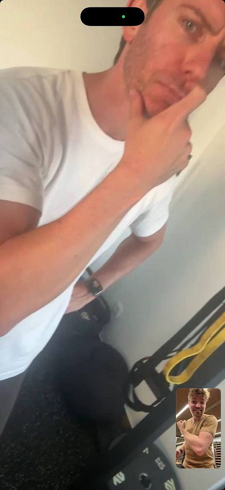 A man in a white t-shirt stands in a gym environment, appearing thoughtful, with fitness equipment and a small inset video call showing another man flexing his arm.