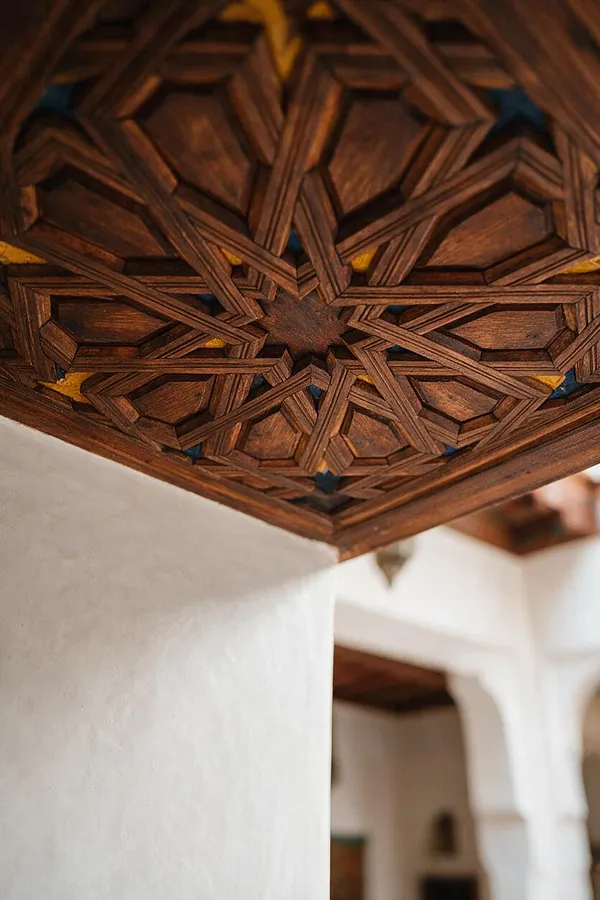 A close-up view of an ornate wooden ceiling featuring intricate geometric carved patterns in a star-like arrangement. Below it are smooth white plaster walls and an arched opening, creating a classic Moroccan interior with warm wood and bright neutral contrasts.