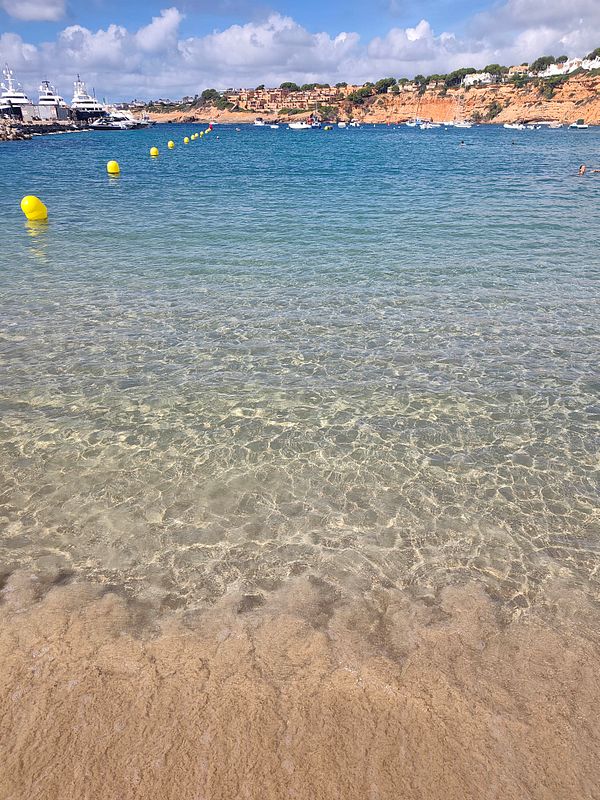 A clear, calm beach scene with transparent shallow water, yellow buoys marking a swimming area, and boats anchored near a coastal town under a partly cloudy sky.