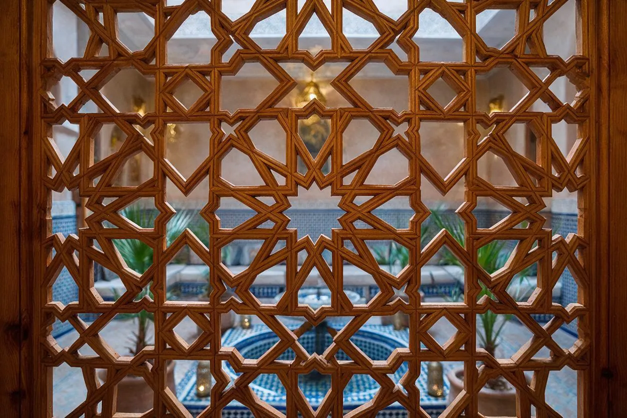 A Moroccan-style exterior view framed by an intricate carved wooden screen with repeating geometric star and diamond patterns. Through the lattice, a blue-tiled courtyard with a small pool, potted palms, and warm stucco walls is visible.