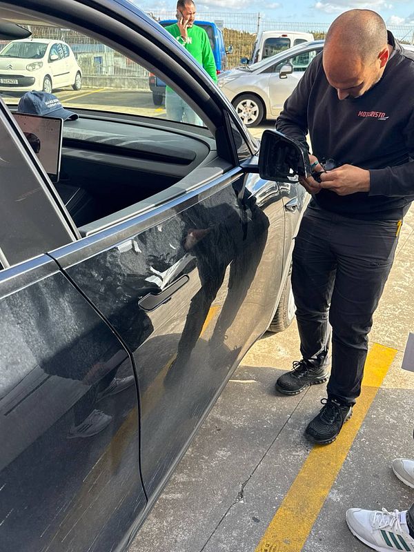 A mechanic is repairing a car's side mirror in a parking lot.