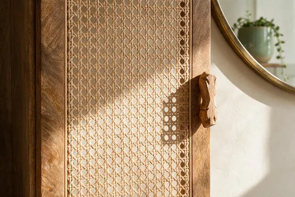 A close-up of a bohemian-style wooden cabinet featuring a woven cane or rattan panel door with a carved wood handle. Soft sunlight highlights the natural texture, while a round mirror and blurred greenery appear in the background.