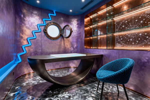 A futuristic home office shows nebula-purple walls with a zigzag electric-blue LED accent, a sculptural gunmetal desk centered on a silver lunar-crater floor, and a peacock-blue woven chair. Bronze-rimmed porthole windows flank copper-backed glass shelving with warm strip lighting and a white star-chart panel, while recessed ceiling spots and a glowing glass desktop reinforce the cosmic, space-inspired atmosphere.