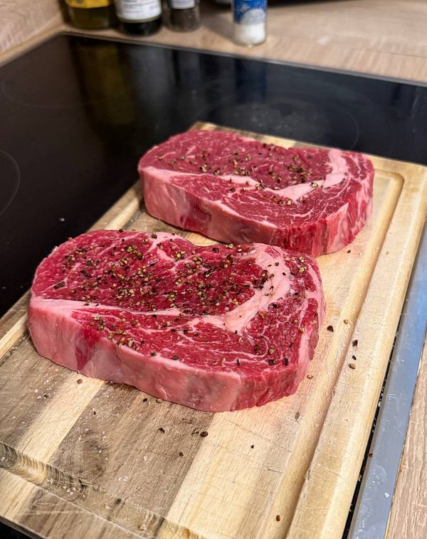 Two raw ribeye steak cuts seasoned with cracked black pepper on a wooden cutting board in a kitchen setting.