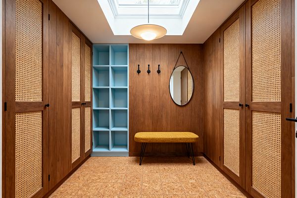 A Mid-Century Modern mudroom with walnut-paneled walls and cane-front closets on both sides, centered by a wall with light blue open cubbies, three matte black hooks, and a round strap-hung mirror. A mustard upholstered bench with black hairpin legs sits below the hooks, cork tile covers the floor, and a large skylight with a saucer pendant floods the space with soft daylight, creating a tidy, symmetrical entry for organized storage.