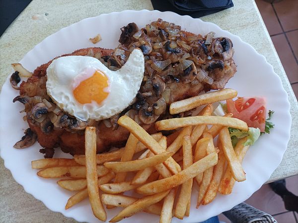 A large plate featuring a schnitzel topped with sautéed onions and a fried egg, accompanied by French fries and a slice of tomato.