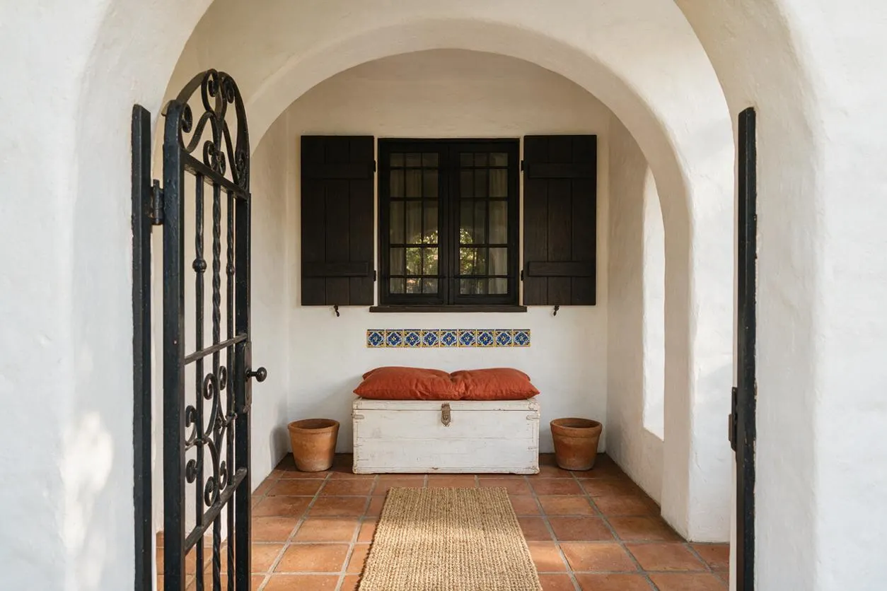 A small Mediterranean-style front porch with white stucco walls, a rounded arch ceiling, terracotta tile flooring, and a black wrought-iron gate. A white wooden chest with rust-colored cushions sits beneath a window with dark shutters, flanked by two terracotta pots and accented by a strip of blue decorative tile.