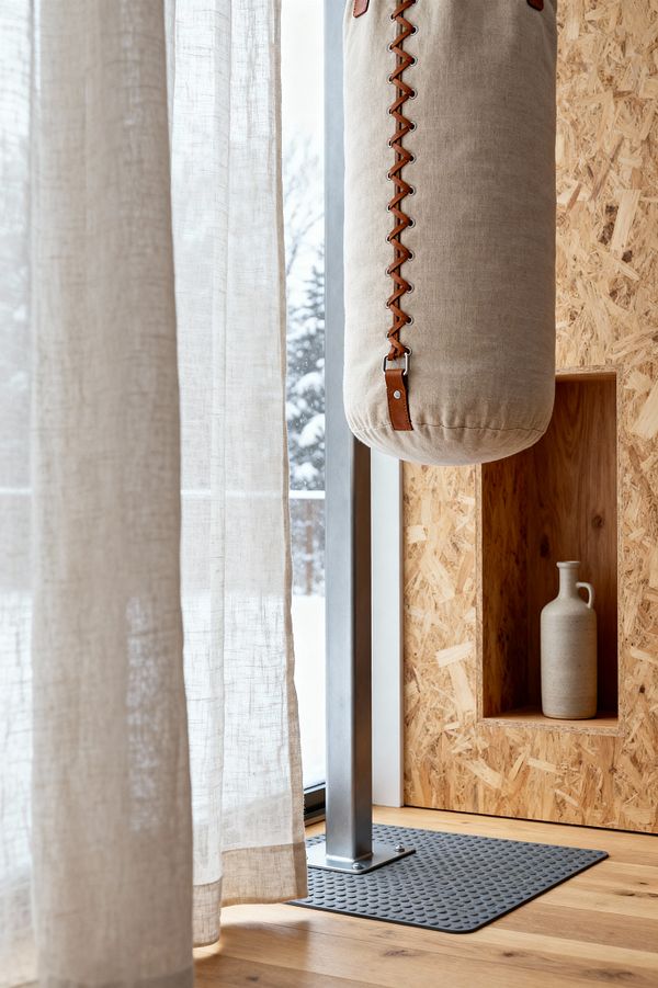 Close-up of a Scandinavian home gym corner with a floor-mounted matte steel stand supporting a beige linen punching bag detailed with tan leather lacing and metal rings, set on a dimpled black rubber mat over light oak flooring. To the right, honey-toned OSB wall cladding includes a recessed niche holding a matte stoneware jug, while to the left sheer off-white linen curtains filter daylight from a tall window with snowy trees visible outside.