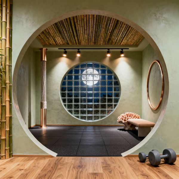 A modern home gym framed by a circular moon‑gate reveals a studio with black rubber floor tiles, sage green plaster walls, and a round glass‑block window centered on the far wall beneath track lights. A rope‑wrapped copper column stands to the left, a copper‑framed round mirror and beige bench sit to the right, and bamboo covers the ceiling and entry, with warm wood flooring and a pair of dumbbells in the foreground.