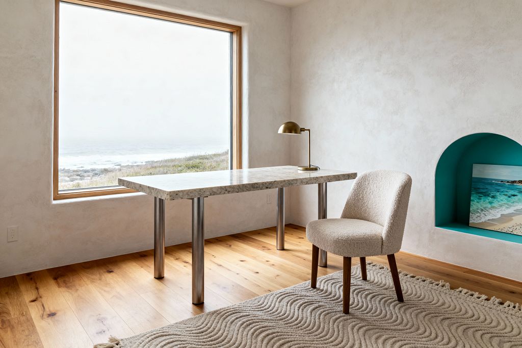 Minimalist home office with a large picture window overlooking a misty coastline. A rectangular desk with a light speckled stone top and cylindrical stainless steel legs sits near the window, topped with a small brass dome lamp. A cream bouclé chair rests on a wave-pattern ivory wool rug over wide-plank oak floors. Warm white plaster walls surround an arched niche painted teal that holds a beach print, creating a calm, sunlit space.