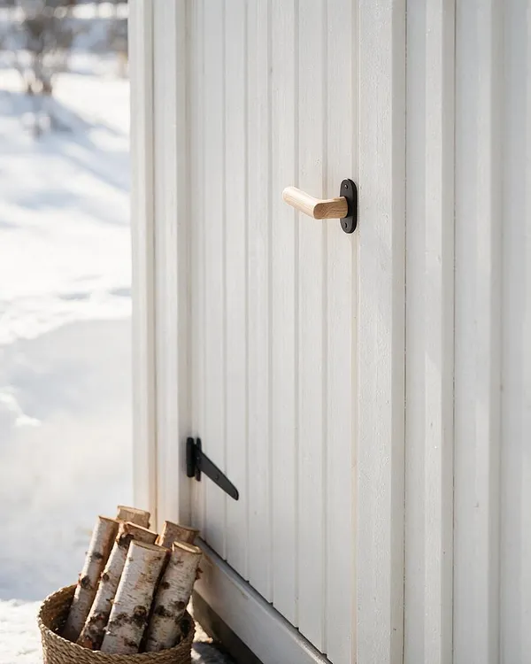 A close-up of a white vertical wood exterior wall or door on a Scandinavian-style cabin, featuring a simple light wood handle and black metal hardware. A woven basket filled with birch logs sits on the snowy ground beside the structure, with soft winter light creating a calm, minimal atmosphere.