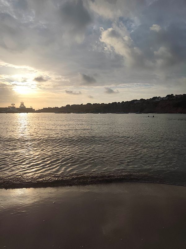 Sunset view of a calm beach with gentle waves and a silhouette of a distant harbor and hills.
