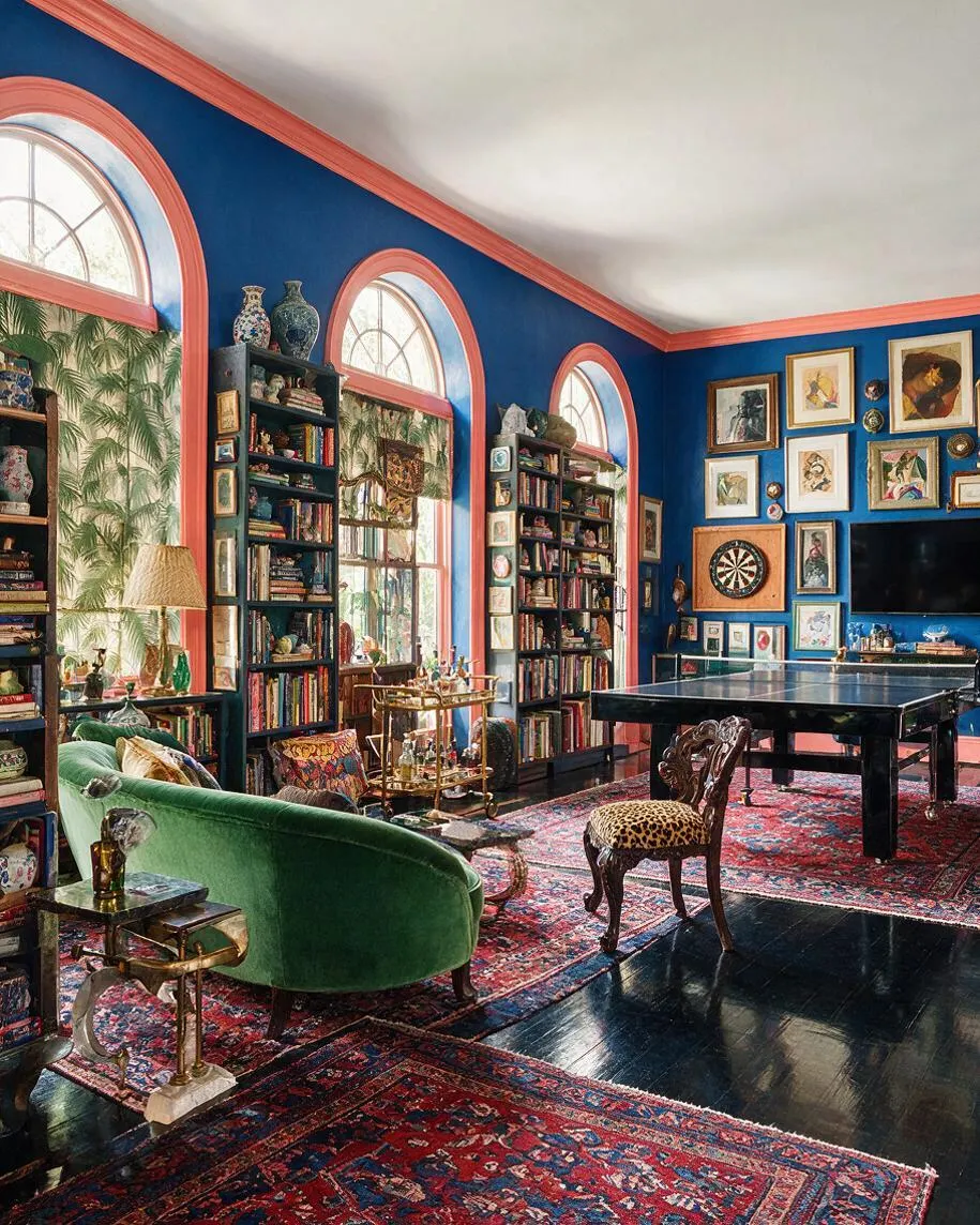A vibrant maximalist game room with deep blue walls, coral trim, and tall arched windows. The space includes a black ping pong table, a green velvet sofa, patterned rugs, dark wood bookcases, and a gallery wall filled with art and decorative objects.