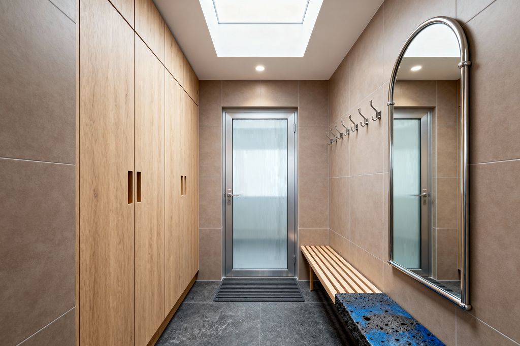 A long, narrow contemporary mudroom features full-height light-oak cabinets on the left and beige tiled walls on the right with a row of metal hooks and a tall arched chrome-framed mirror. A slatted wood bench and a charcoal terrazzo bench with blue flecks sit on dark gray stone-look tile flooring, while a central skylight and recessed lights brighten frosted reeded-glass doors framed in aluminum.