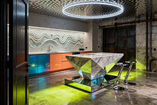 An industrial-chic kitchen features a faceted white marble island on a mirrored plinth, lit from below with neon yellow-green. Glossy teal and orange cabinets line the back wall beneath a sculpted, wavy pale seafoam ceramic backsplash. Overhead, a large LED ring floats against a perforated metal ceiling. Polished concrete floors, raw plaster walls, a weathered steel door, and two chrome Z-shaped stools complete the psychedelic, high-contrast scene.