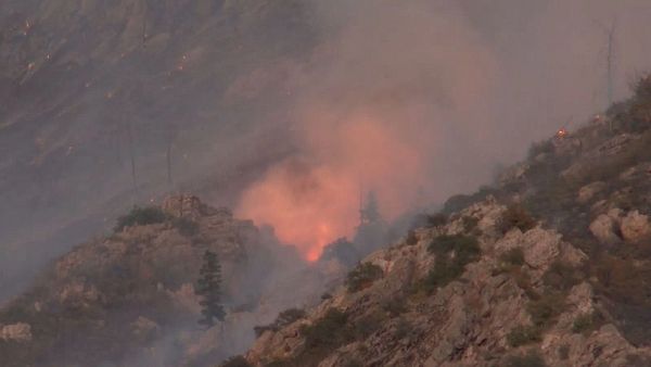 A wildfire is actively burning on a mountainous terrain, surrounded by smoke.