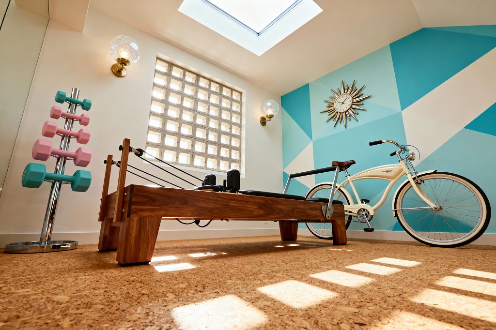 Bright home gym with a wooden Pilates reformer sits on cork flooring beneath a large skylight. A teal and aqua geometric mural with a brass sunburst clock spans one wall, while brass-and-glass globe sconces flank a square glass-block window. A chrome dumbbell tree holds pastel pink and turquoise weights, and a cream cruiser bicycle rests nearby, creating a clean, retro atmosphere with natural wood, warm light, and crisp lines.