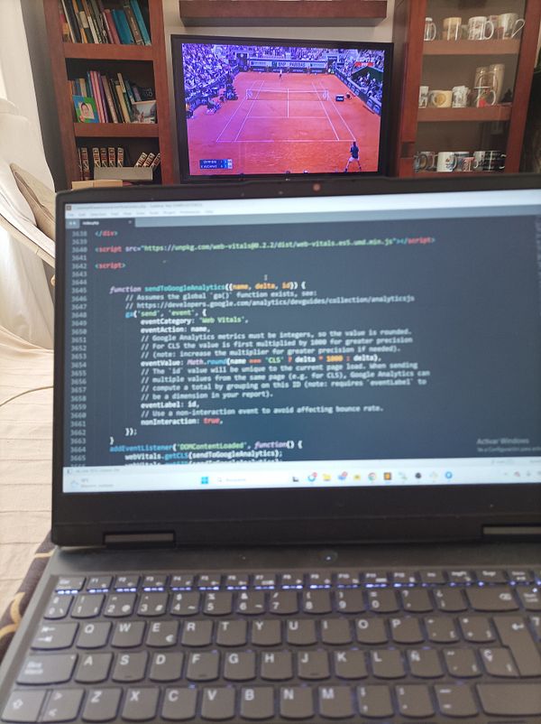 A laptop displaying code is positioned in front of a television showing a tennis match at Roland Garros.
