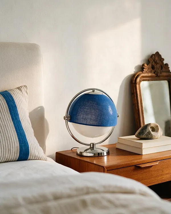 A cozy bedroom vignette shows a cream upholstered headboard and bed in the foreground, a striped pillow with a bold blue band, and a warm wooden nightstand. On the nightstand sits a blue-and-chrome globe-style lamp, with stacked books and a small stone object beside an ornate vintage mirror leaning against a white wall.