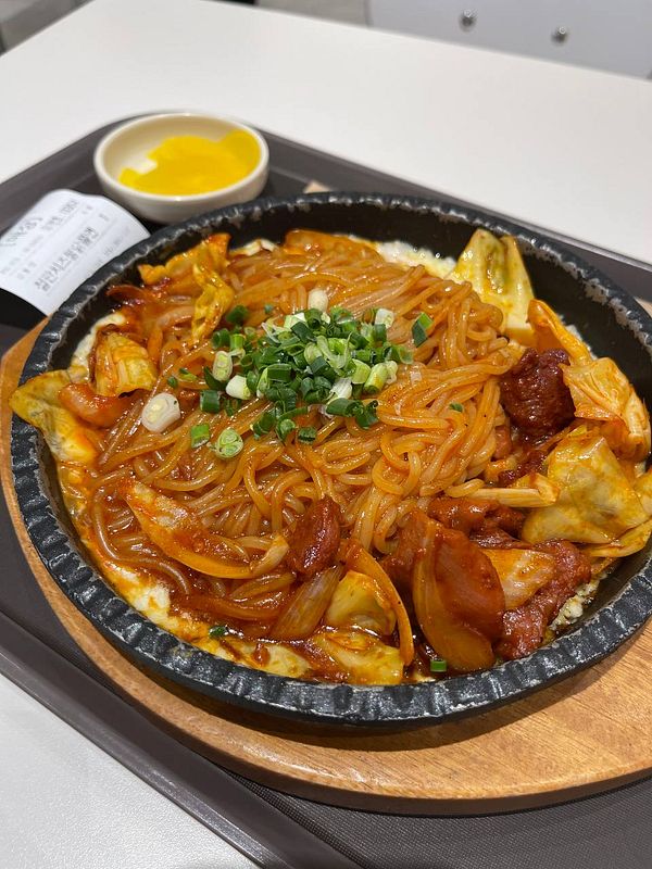 A sizzling plate of stir-fried noodles topped with vegetables and garnished with green onions.