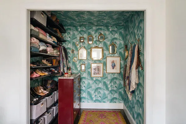 A narrow closet styled in a maximalist way with teal palm-print wallpaper covering the walls and ceiling. Open shelves on the left hold folded clothes, shoes, and storage baskets, while the right wall displays hanging scarves and accessories. A small red cabinet with brass hardware and several gold-framed mirrors and pictures add a layered, decorative look.