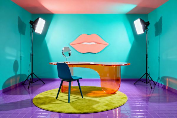 A pop art home office features turquoise walls and a coral pink ceiling with a large peach-colored lips wall sculpture centered behind a translucent orange acrylic desk. The desk sits on a circular lime-green rug over glossy purple tiles and is paired with a teal chair and a chrome round tabletop mirror; two studio softbox lights on black tripods flank the setup, creating even light that highlights bold shapes and saturated colors.