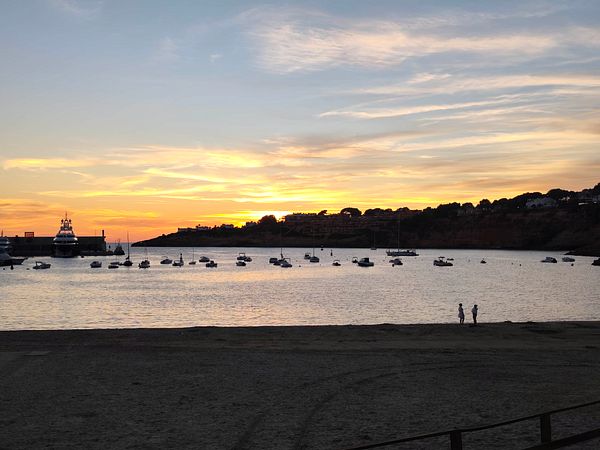 Sunset view of a calm bay with multiple boats anchored and two people standing on the beach.