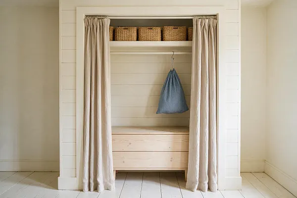 A simple coastal-style closet nook with cream linen curtains framing an open alcove. Inside are woven basket storage bins on the top shelf, a single hanging blue fabric bag, and a pale wood bench or chest below, all against white shiplap walls and light painted floors.