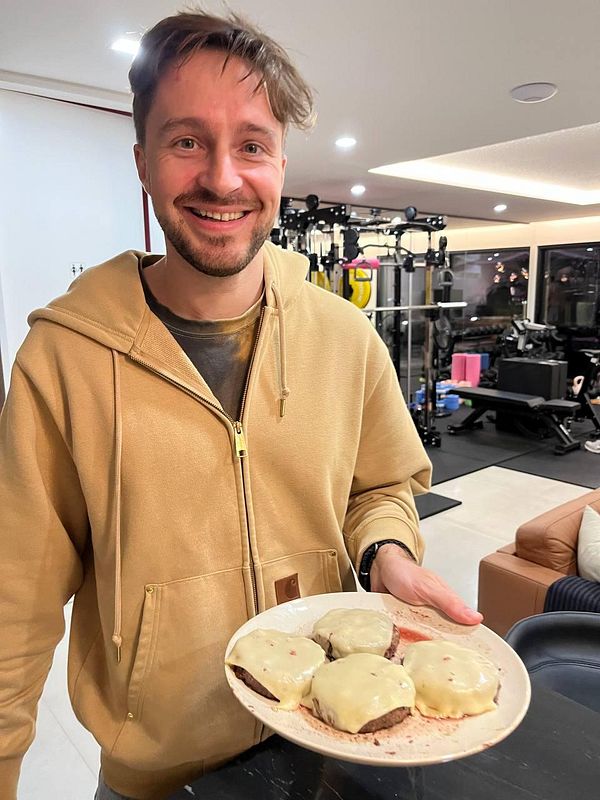 A smiling man holds a plate of cheeseburgers in a modern indoor setting.