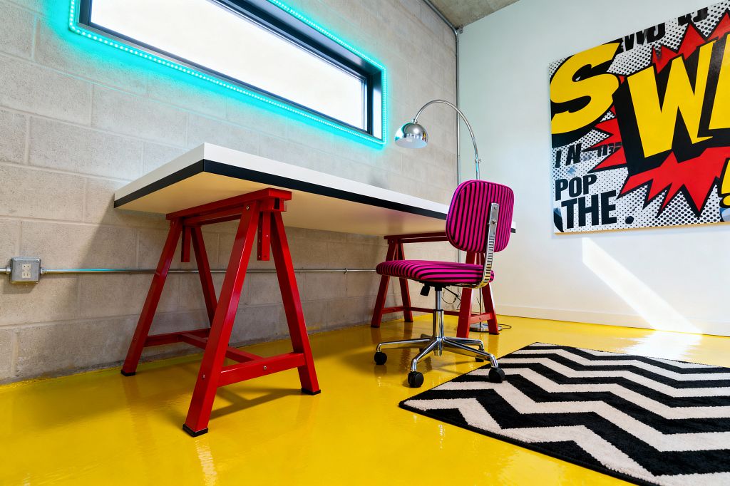 A pop art–inspired home office features a high-gloss yellow floor, a black-and-white chevron rug, and a white laminate desk with black edge banding supported by red trestle legs against a light concrete-block wall. A magenta-and-black striped swivel chair with a chrome base sits beside a chrome task lamp, a narrow window rimmed with teal LED lights, and a large comic-style canvas in bold yellow, red, black, and white for an energetic, industrial feel.