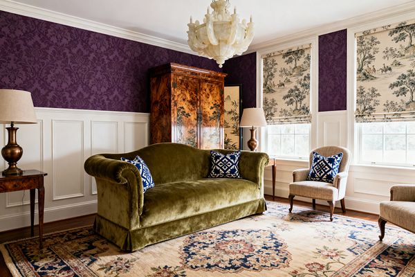 Traditional media room with deep plum damask wallpaper above white wainscoting, centered by an olive green velvet roll‑arm sofa with blue-and-white pillows. Beige upholstered armchairs flank a floral wool rug, while three windows with cream toile Roman shades bring light; a sculptural cream chandelier, bronze table lamps, and a glossy burl‑wood armoire add classic warmth.