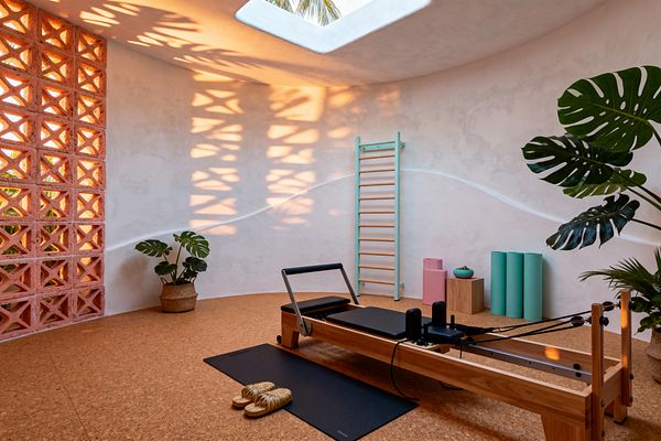 Sunlit home gym with a wood Pilates reformer on cork flooring sits below a rectangular skylight where palm fronds are visible, while a coral terracotta breeze-block wall casts patterned shadows on warm white plaster. Mint wall bars, seafoam rollers, a pastel pink block, a small wood pedestal, a black mat with straw slippers, and potted monstera and palm plants complete the space.