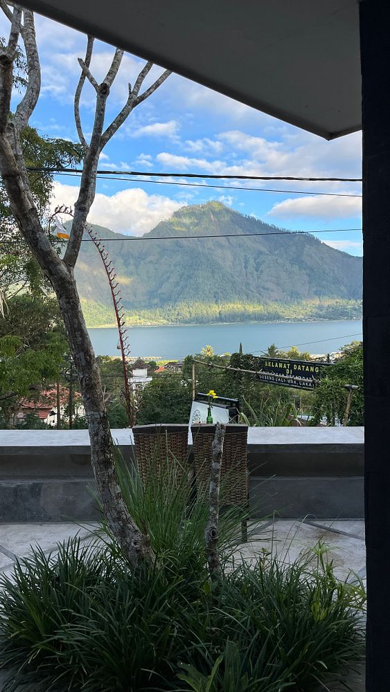 A scenic view of Kintamani featuring a mountain and lake, framed by greenery and a modern outdoor seating area.