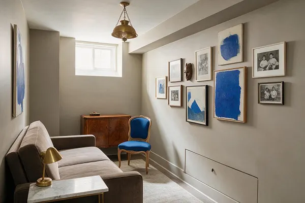 A narrow studio apartment sitting area with beige walls, a small high window, and a brass pendant light. A taupe sofa with a marble-topped side table and brass lamp faces a blue upholstered accent chair, a wood cabinet, and a gallery wall of blue abstract art, black-and-white photos, and a carved mask.