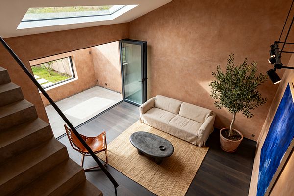 A high-ceilinged contemporary living room with warm terracotta plaster walls, a large roof skylight, and black-framed folding glass doors opening to a small courtyard. Dark wood floors support a jute rug, beige linen sofa, oval charcoal concrete coffee table, and a cognac leather sling chair. A potted olive tree in a terracotta planter sits beside a bold blue canvas, while black track spotlights and a minimalist stair with a dark metal rail complete the space.