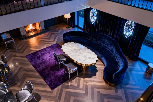 A double-height living room features a sweeping sapphire velvet curved sofa around a scalloped white marble coffee table with a polished brass base, set on a rich purple rug over herringbone wood floors. Floor-to-ceiling dark violet velvet drapes with two faceted, diamond-like sconces, a marble-surround fireplace, a brass floor lamp, and chrome-framed silver chairs complete the moody, jewel-toned Vintage Glam scheme.