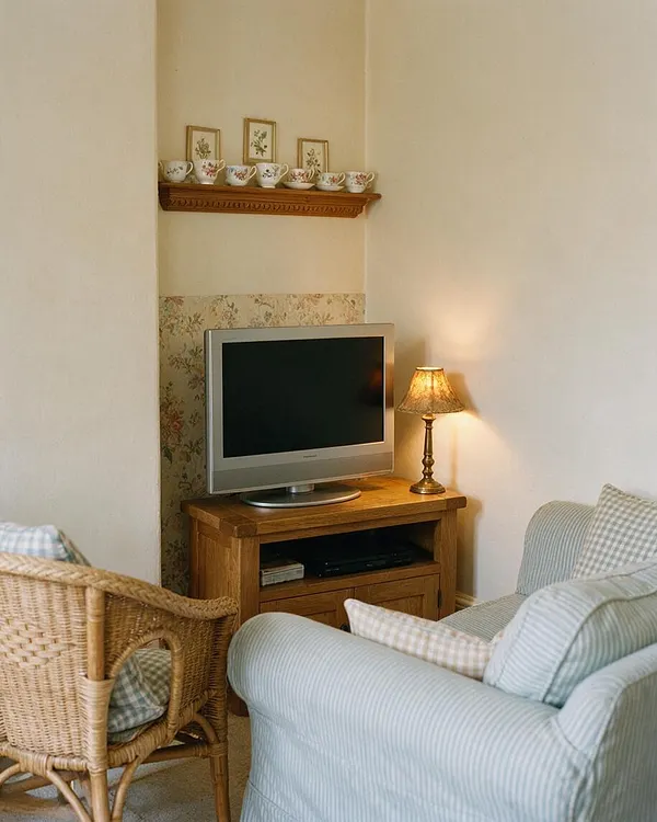 A small cottagecore media room with a silver television on a wooden stand, a glowing table lamp, and a wicker chair in the foreground. Soft striped upholstered seating and a floral wallpaper accent panel create a warm, nostalgic atmosphere, with a shelf above holding decorative teacups and botanical prints.