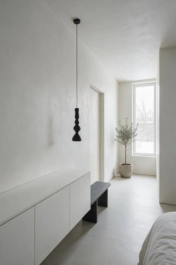 A bright minimalist sunroom with smooth white walls, a tall narrow window, and a potted olive tree in a light stone planter. The space features a long low white cabinet, a small dark bench, and a sculptural black pendant light hanging from the ceiling.