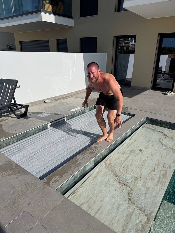 A man is playfully balancing on a pool cover while standing barefoot in a sunny outdoor setting.