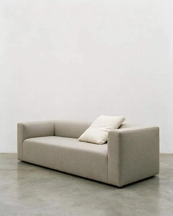 A low, boxy beige sofa sits against a plain white wall on a polished concrete floor. It has straight arms, a smooth upholstered finish, and a single off-white cushion leaning on the right side, creating a simple minimalist look.