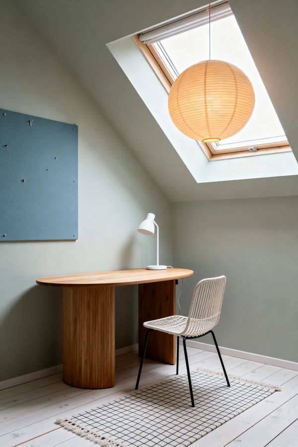 A compact Scandinavian home office under a sloped ceiling with a skylight shows an oval oak desk with fluted pedestal bases, a white metal task lamp, and a white woven rattan chair on black legs over a cream rug with a black grid. Soft sage walls hold a blue felt pinboard, while a spherical rice paper pendant warms the space; pale whitewashed floorboards and minimal decor create an airy, calm workspace.
